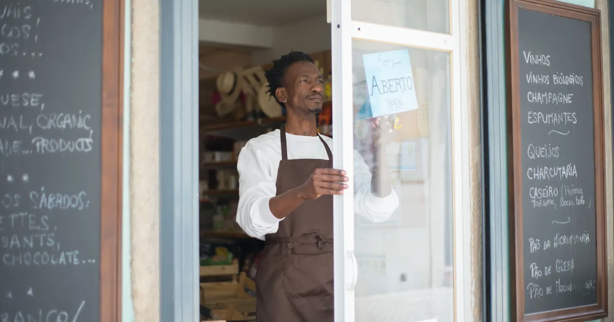 Commerçant en tablier marron qui ouvre la porte vitrée de son épicerie de quartier, avec des ardoises de produits de chaque côté