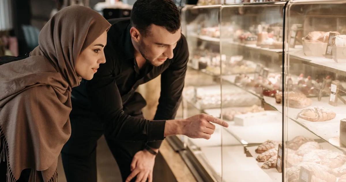 Deux clients penchés devant la vitrine réfrigérée d'une pâtisserie, l'un montre une pièce du doigt