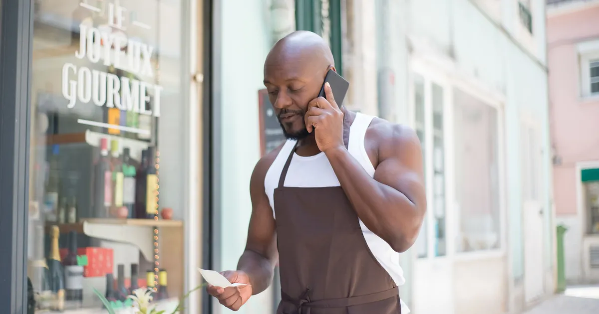 Commerçant en tablier qui téléphone devant sa vitrine d'épicerie fine en lisant une note