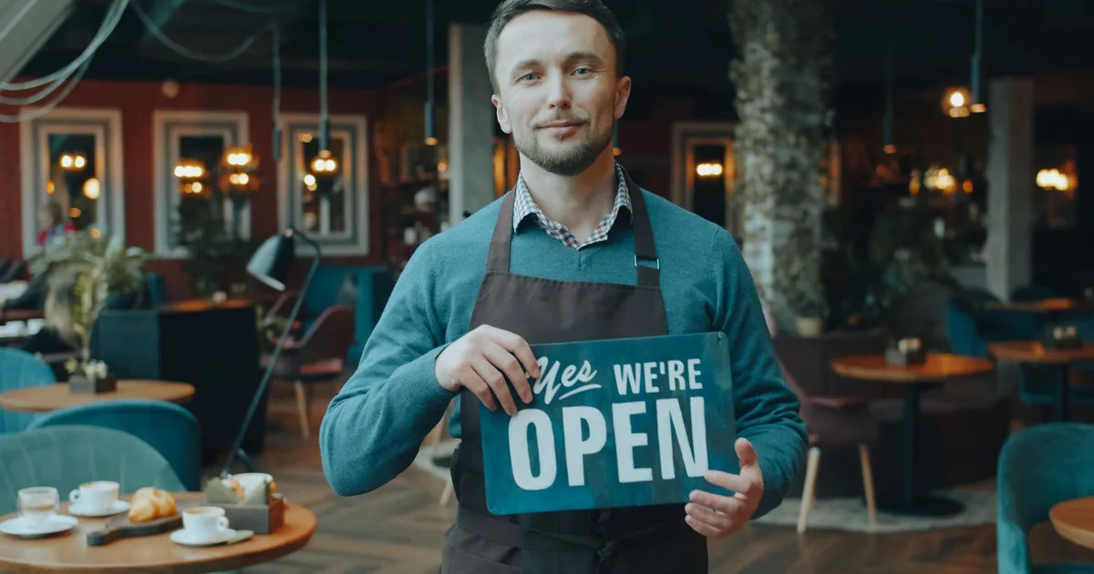 Smiling shop owner in an apron holding a Yes We Are Open sign inside a cafe