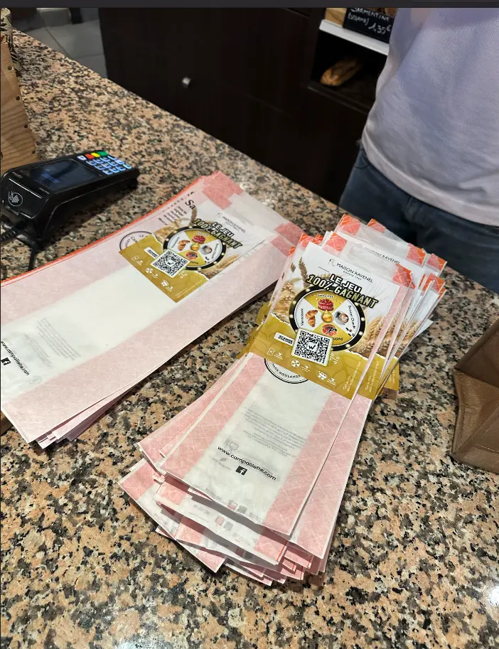 Sachets de baguette avec le flyer du jeu concours Maison Ravenel agrafé, empilés sur le comptoir